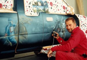 Punaisiin pukeutunut Simo Riikonen kyykyssä henkilöauton vieressä, jonka siniseen kylkeen hän maalaa ratsastavia ritareita.