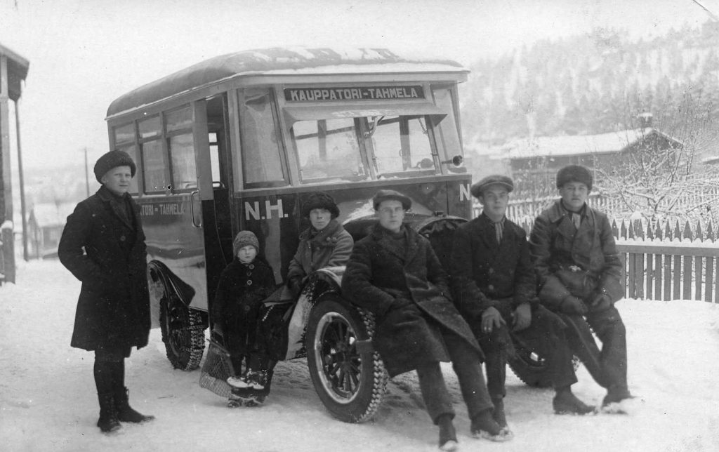 Mustavalkokuvassa miehiä istumassa nokkamallisen linja-auton etupuskurilla.