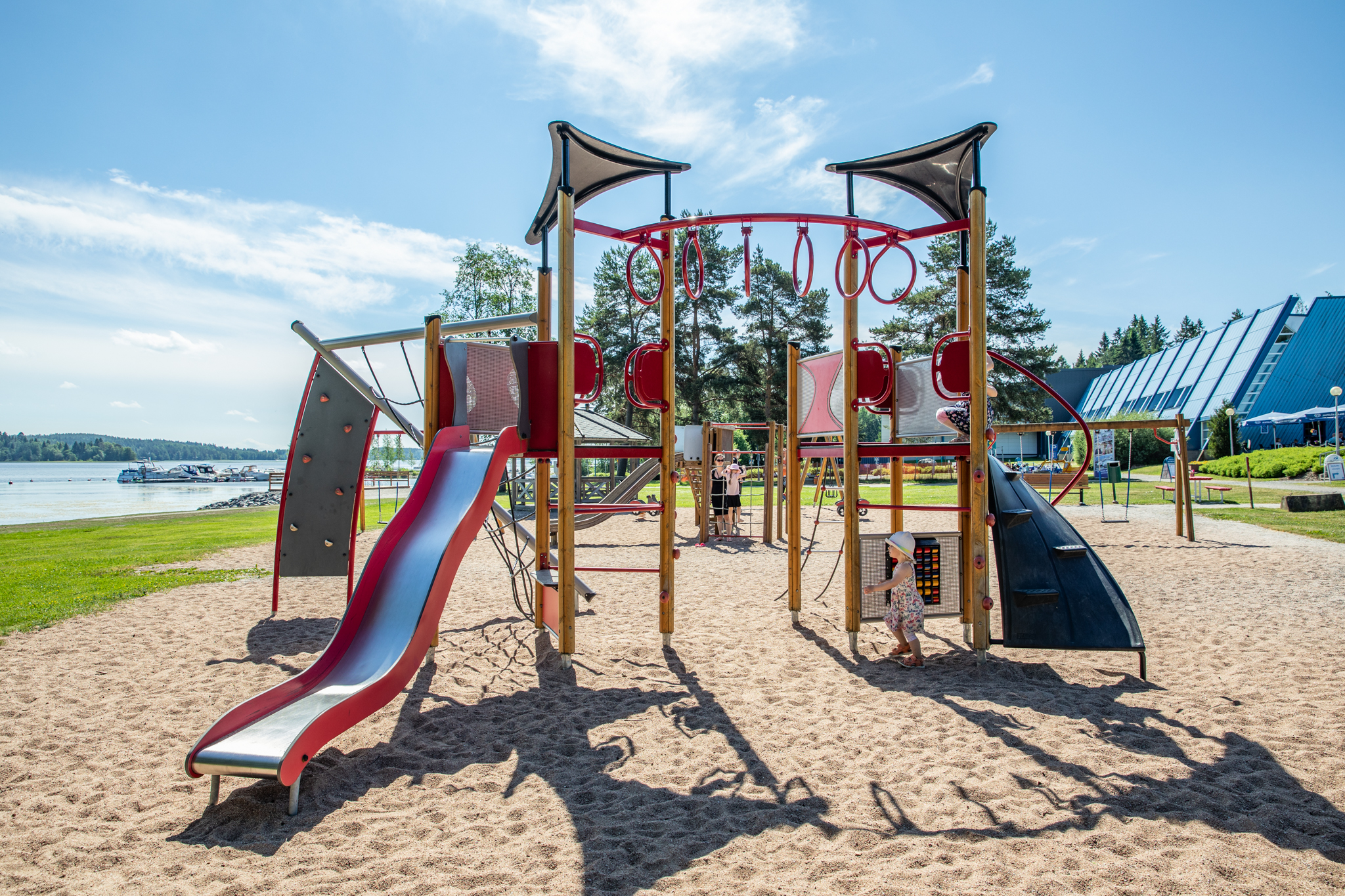 Aurinoinen lasten leikkipuisto kiipeilytelineineen ja liukumäkineen ranta-alueella.