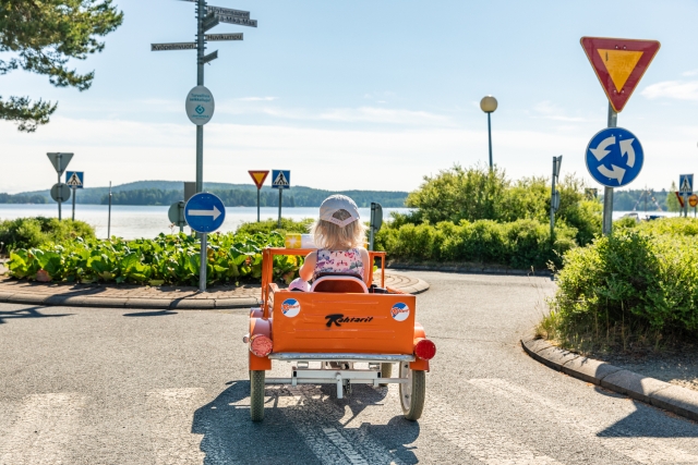 Pieni tyttö ajaa oranssilla polkuautolla kiertoliittymään rannalla sijatisevassa liikennepuistossa.