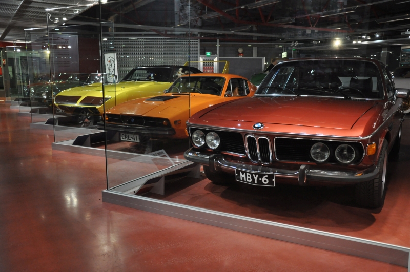 Värikäs rivi urheilullisia menopelejä lasiseinän takana: edessä viininpunainen BMW, sen takana oranssi Saab ja keltainen Plymouth.