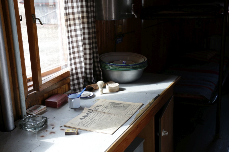 Ikkunan edessä olevalla pöydällä on päivän Aamulehti, partasuti, pesuvateja sekä tuhkakuppi. Aika on pysähtynyt ruudullisten verhojen taakse asuntoparakkiin.