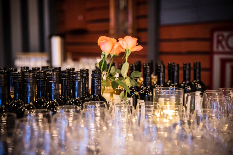 Viinilasien ja viinipullojen rivistö juhlakattauksessa oranssipunaisten ruusujen edessä.