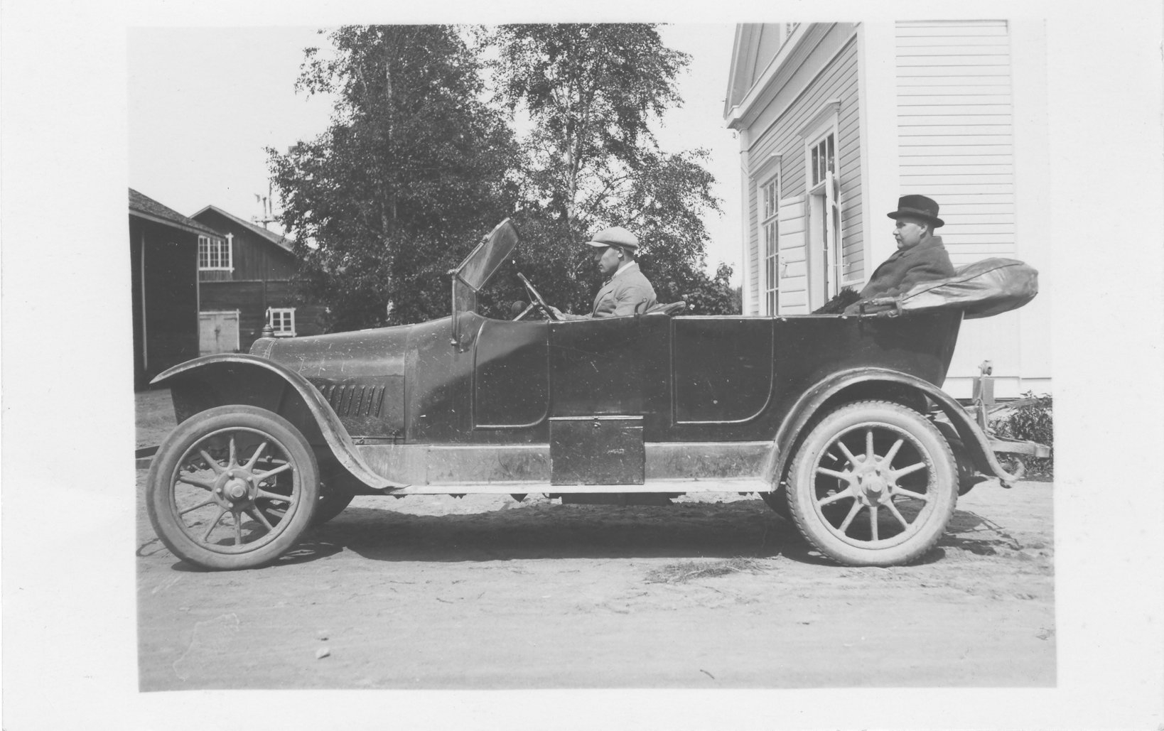 Vanhassa mustavalkokuvassa pinnapyöräinen, tumma Opel, jonka ratin takana istuu kuljettaja ja takapenkillä kuomu laskettuna tyylikäs, hattupäinen mies.