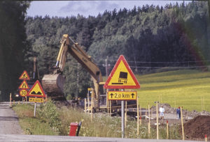 Tietyömaa maalaismaisemassa. Monta erilaista eri vaaroista varoittavaa liikennemerkkiä tien reunassa ja keltainen kaivinkone niiden takana rinteessä työssä.