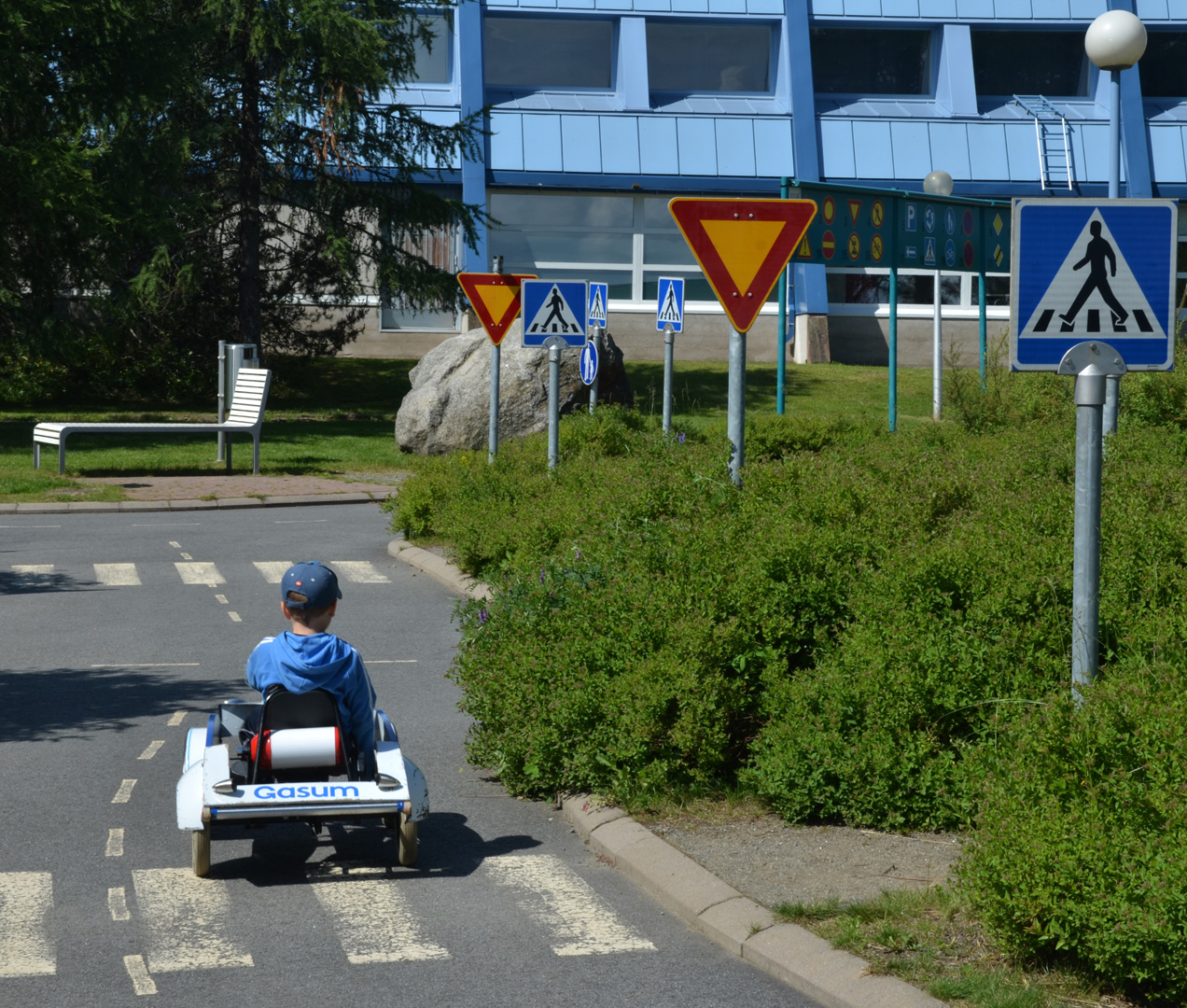 sinivaatteinen lapsi ajaa valkoisella polkuautolla poispäin kuvaajasta liikennepuiston tiellä isojen pensaiden ohi