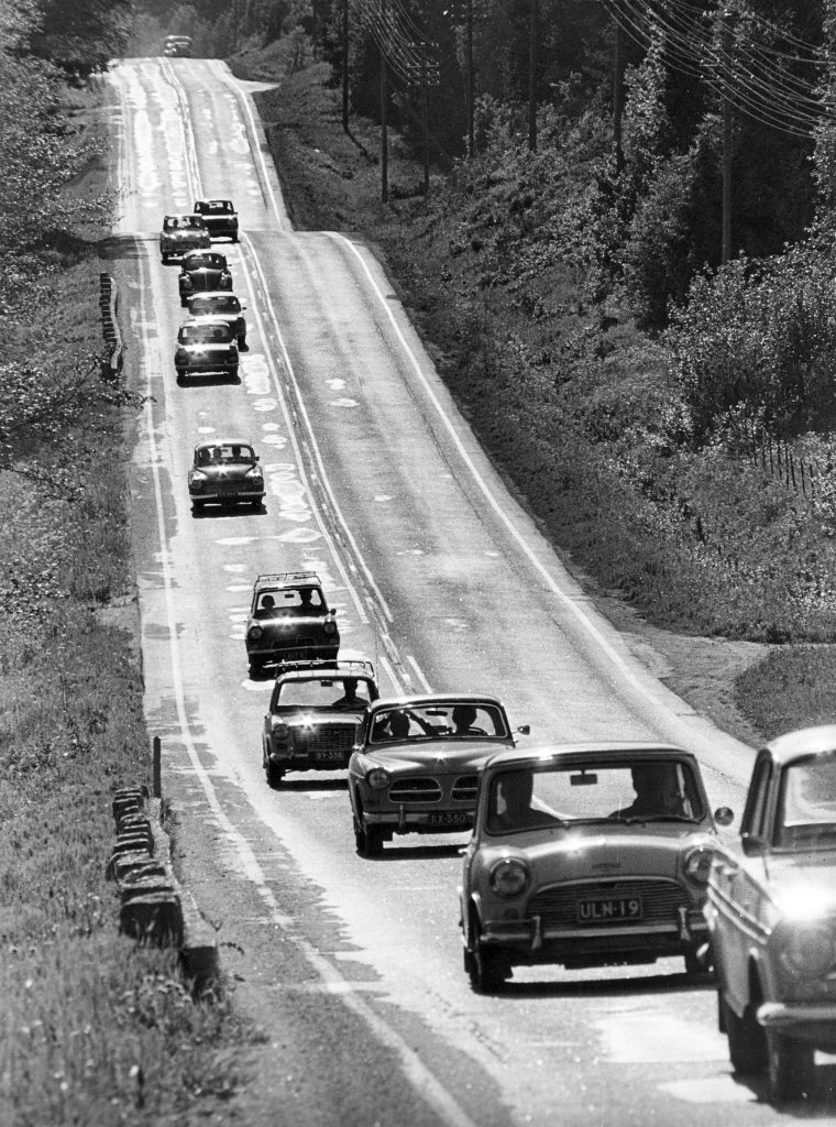 Mustavalkoisessa kuvassa juhannukselta 1969 autoletka aikakauden henkilöautoja etenee kohti kuvaajaa kumpuilevalla päällystetiellä. Vastakkainen kaista on tyhjä yhtä horisonttiin häviävää autoa lukuunottamatta.