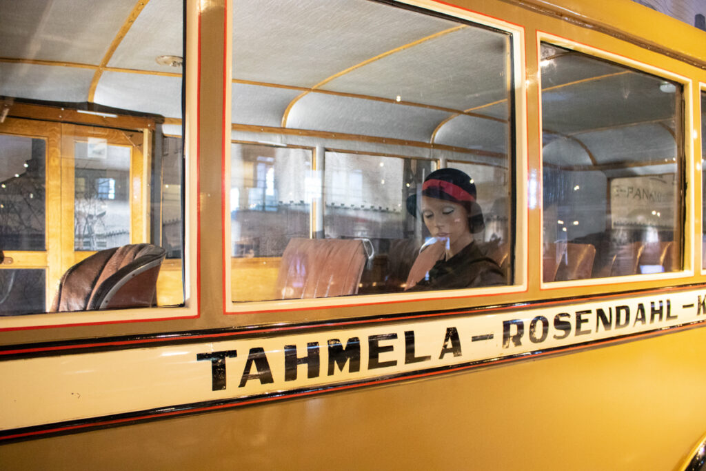 Kellertävän ruskea vanhan linja-auton kylki, jossa lukeea Tahmela-Rosendahl. Linja-auton ikkunassa istuu matkustajaa esittävä naisnukke hattu päässään.
