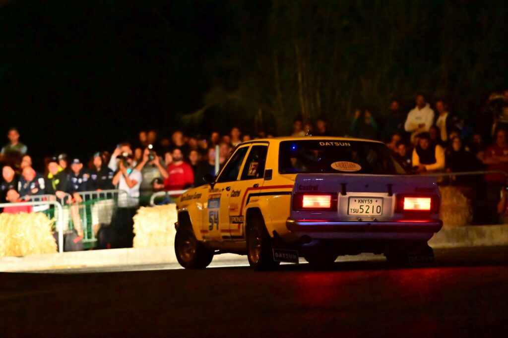 Pimeässä illassa Datsun-historicralliauto ajamassa pois päin katsojasta kohti valokuvaajien muuria.