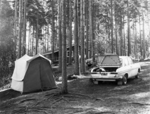 Mustavalkokuvassa näkyy mäntypuiden takaa pieni mökki. Kuvan etuosassa puiden lomaan pystytetty teltta ja henkilöauto, jonka takakontin luukku on auki.