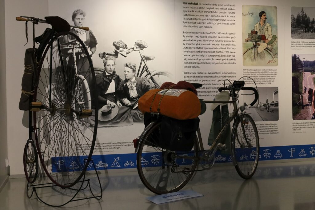 Näyttelyn tekstiseinän edessä on isolla etupyörällä varustettu isopyöräinen sekä sen vieressä nykyaikaisempi polkupyörä. Molemmat on pakattu matkapakaasein pyörällä tehtävää matkaa varten.