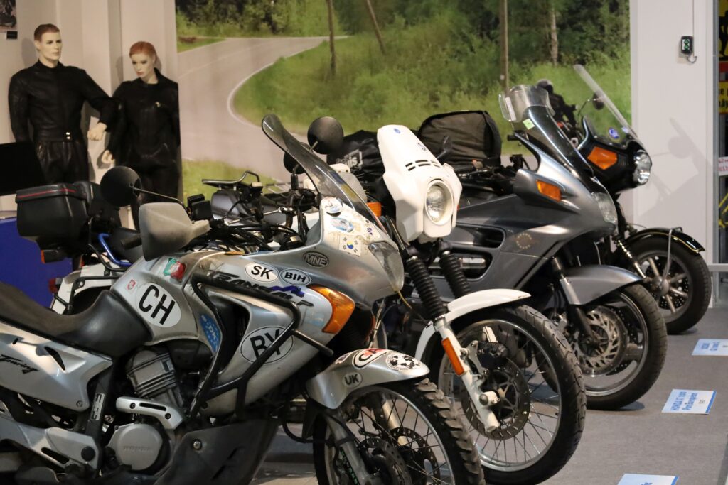 Näyttelyn moottoripyörärivistö, jonka takana kaksi motoristin ajopukuun puettua nukkea ja suurkuva metsäiseen maisemaan kaartuvasta asfalttitiestä.