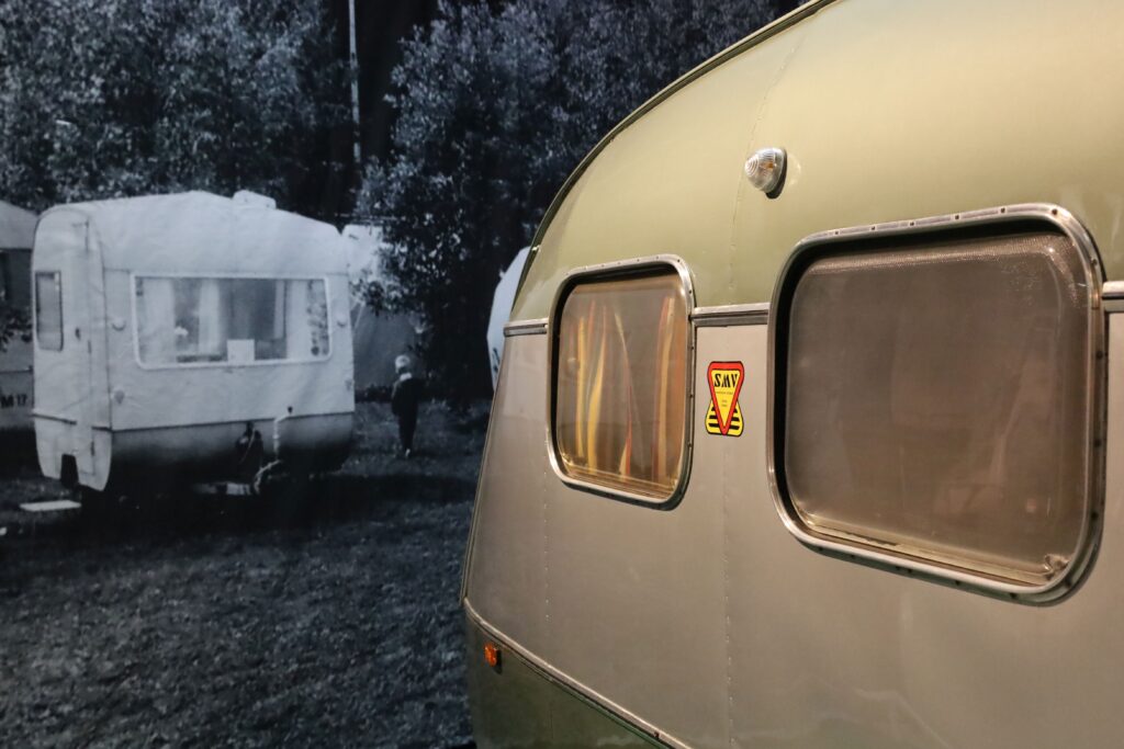 SMW-merkkisen 1970-luvun matkailuvaunun kaareva takaosa jatkaa vaunun takana olevassa mustavalkoisessa kuvassa olevaa leirintäalue teemaa.