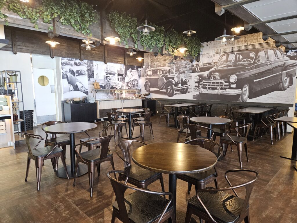 Tummasävyinen kahvila, jossa on tummaa puuta olevat pyöreät pöydät ja seinissä isot mustavalkoiset kuvat ajoneuvoista.
