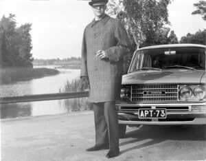 Mustavalkoisessa kuvassa mies seisoo tupakka kädessään Toyotan keulan edessä taustalla järvimaisema tien vieressä.