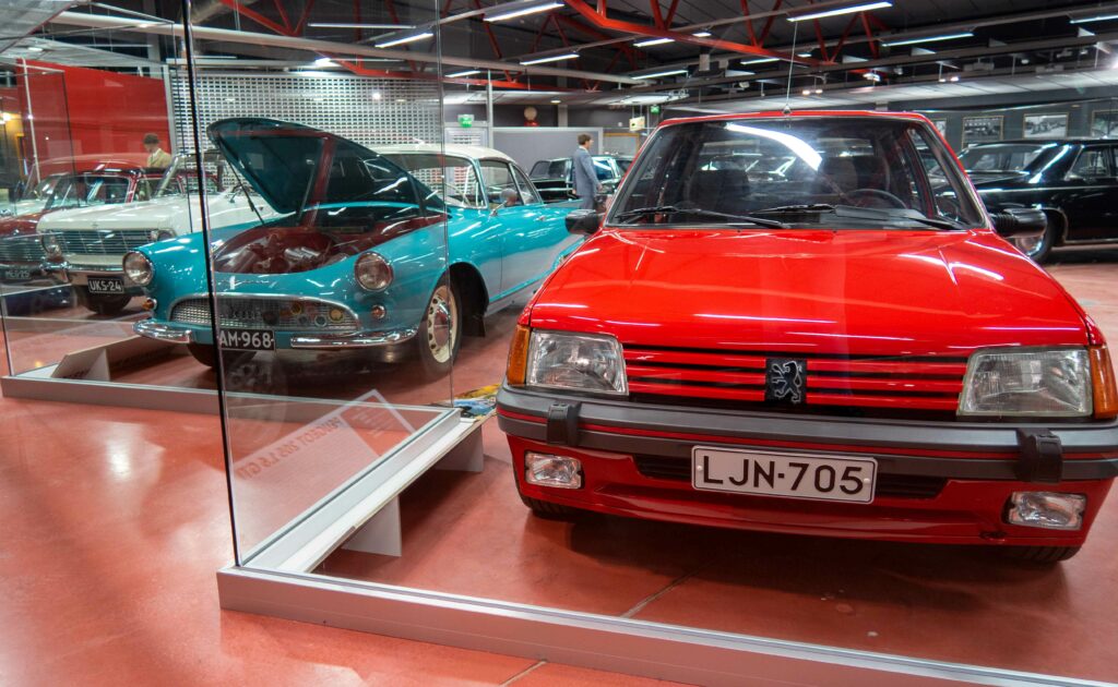 Lasiseinän takana 80-luvun kirkkaanpunainen Peugeot 205 GTI ja sen takana vasemmalla näkyy turkoosin sininen Autounion 1000 SP coupe 60-luvulta konepelti avoinna.