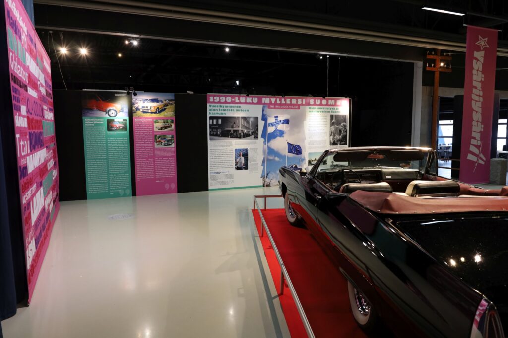 Musta avo-Cadillac kuvattuna takaviistosta näyttelytilassa punaisella matolla. Taustalla näkyy 90-luvun pinkissä ja mintunvihreässä värissä olevia näyttelyseinäkkeitä.