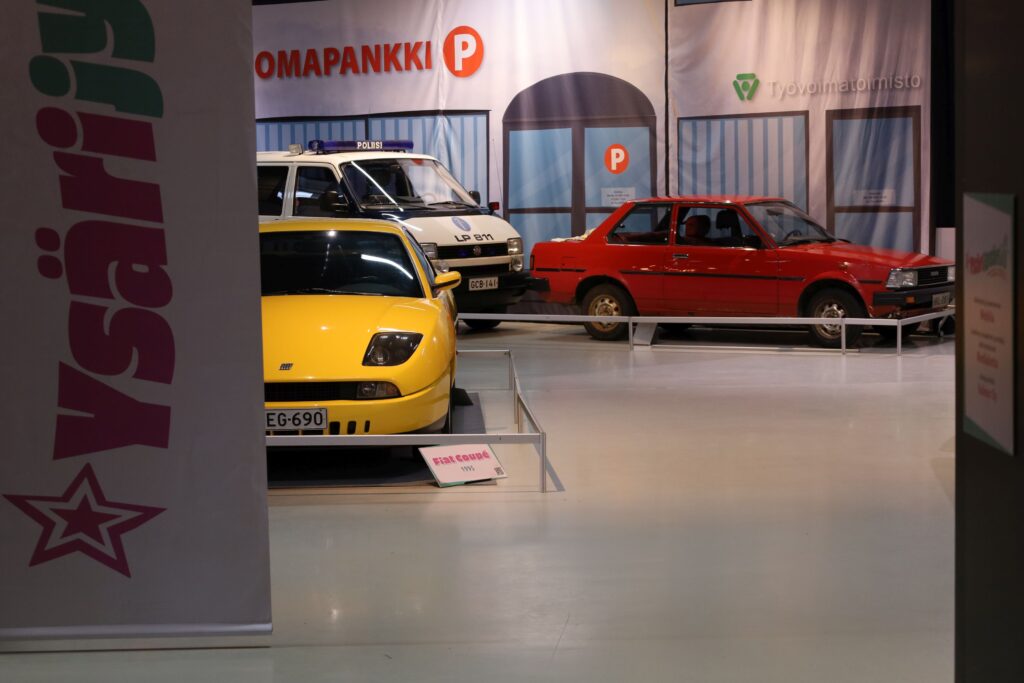 Näyttelyn sisään käynti, reunassä Ysärijysäri-logo pystysuorassa kankaassa, sen takana kirkkaankeltainen Fiat sekä taustalla punainen Corolla poliisi-Maija perässään katumaisemassa.