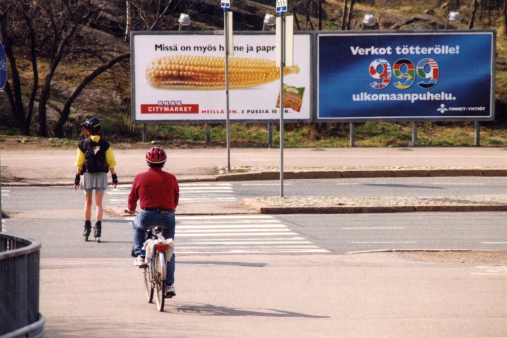 1990-luvun värikuvassa rulluluistelija ja polkupyöräilijä lähestyvät kevyenliikenteen väylällä ajotien ylittävää suojatietä. Toisella puolella tietä näkyy isot mainoskyltit, joista toinen mainostaa Citymarketin herne-maissi-paprikaa ja toinen Finnetin 999-ulkomaanpuheluita.