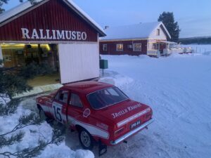 Punainen Ford Escort Twin Cam -ralliauto vm. 1968 talvisessa pakkassäässä odottamassa siirtoa sisään Rallimuseon näyttelytilana toimivaan punaiseen puurakenteiseen ulkorakennukseen. Taustalla näkyy Sevetin baarin luminen katto.
