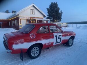 Puna-valkoinen Ford Escort Twin Cam -ralliauto vm. 1968 sivulta kuvattuna lumisessa maisemassa puna-valkoisen puurakkennuksen edessä.