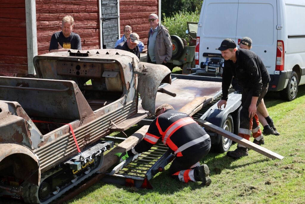 Kauniina kesäpäivänä ulkona nurmikolla otettu kuva, jossa huonokuntoista vanhaa ajoneuvon koria siirretään apuvälinein trailerille. Ympärillä useita ihmisiä seuraamassa toimenpidettä.