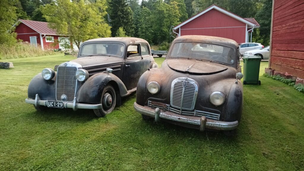 Nurmikolla kuvattu edestäpäin kaksi hyvin pölyistä vanhaa autoa Mercedes-Benz 170 S ja Austin A70 Hereford.