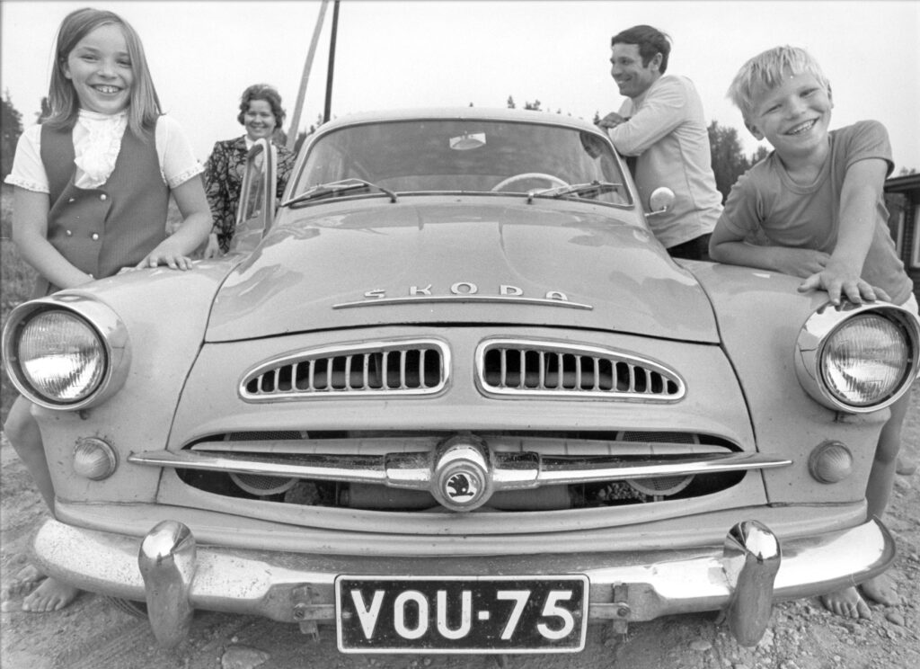 Mustavalkoinen kuva vuodelta 1971, jossa kuvan keskellä keulasta kuvattu Skoda-henkilöauto, jonka toisella puolella autoon nojaa iloisesti kameraan hymyillen tyttö ja toisella poika. Perheen äiti ja isä näkyvät auton vieressä taustalla.